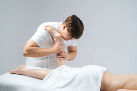 Male masseur with strong hands massaging lower part of leg to young woman lying on massage table on white background. Professional therapist men doing wellness massage shins and legs for female clientの写真素材