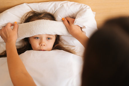 Close-up of mother putting compress on forehead to bring down temperature of sick daughter with fever lying under blanket at home. Parent caring for sick child lying in bed with flu.の写真素材