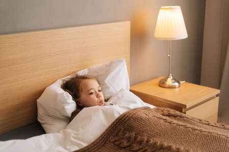 Side view of adorable little sick girl lying in bed measuring temperature with thermometer in mouth at home. Upset child with high fever lying at bad in medical clinic. Concept of children health.の写真素材