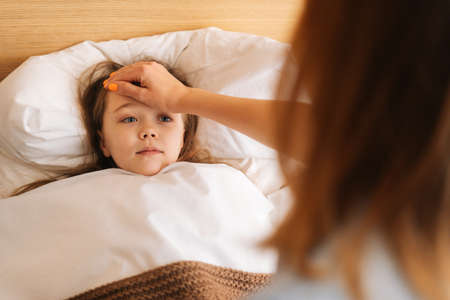 Close-up view of young mother measures temperature touches forehead of sick daughter lying in bed at home. Parent caring for sick child lying in bed with flu. Concept of children health.の写真素材