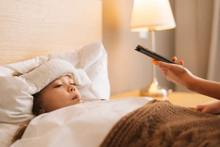 Close-up of worried mother sitting on bed beside sick daughter with high fever and calling on phone to family doctor. Parent caring for sick child lying in bed with flu. Concept of unwell child.の写真素材