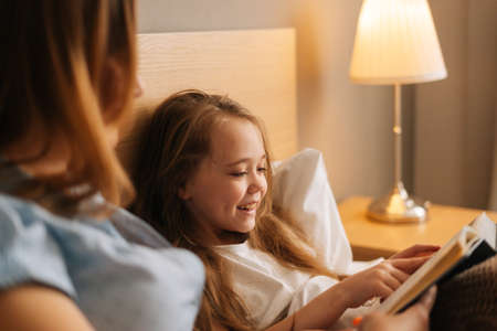 Close-up of mother and cheerful adorable daughter reading together children book before going to sleep while lying in bed in nursery bedroom, near lamp. Concept of family leisure activity at home.の写真素材