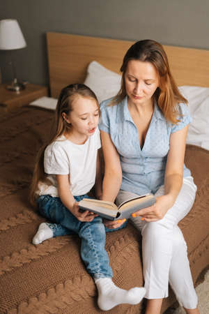 Portrait of young mother and adorable daughter reading children book together sitting on bed in nursery bedroom. Concept of family leisure activity at home.の写真素材