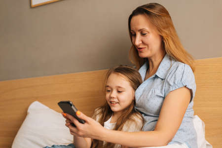 Happy young mother hugging to preschool daughter together browsing and surfing internet on mobile phone, chatting on video call sitting on bed in bedroom. Concept of family leisure activity at home.の写真素材
