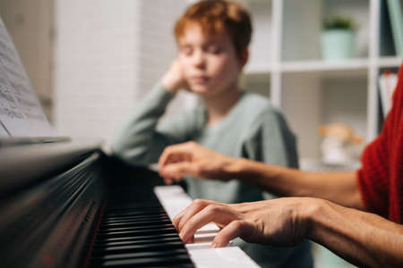 Focused redhead child student boy listening his teacher play the piano during lesson. Father teaching son to play musical instrument in living room. Concept of music education.の写真素材