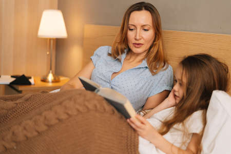 Close-up view of young happy mother and adorable cheerful daughter reading children book together lying in bed before going to sleep. Concept of family leisure activity at home.の写真素材