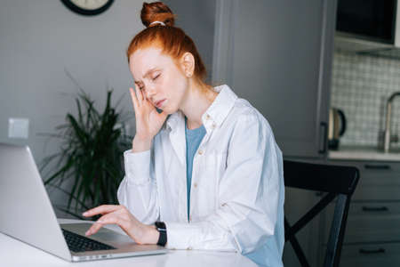 Suffering redhead young woman massaging temples, reducing painful head feelings during work on laptop computer at home workplace. Concept of remote working from home office.の写真素材