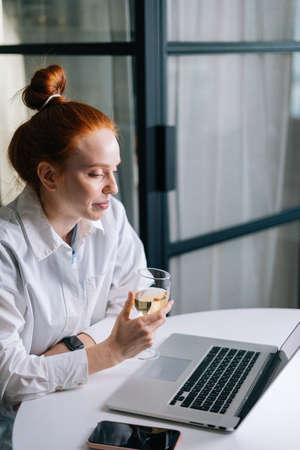 Beautiful redhead young woman having video call conversation using laptop and drinking champagne. Happy lady talking via webcam gathering friends, boyfriend sending online congratulations.の写真素材