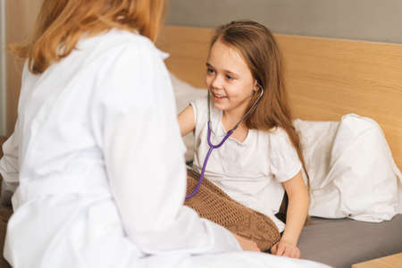 Portrait of cheerful little girl listening heartbeat with stethoscope to female doctor sitting in bed. Child playing as doctor nurse listening physician with stethoscope. Concept of children health.の写真素材