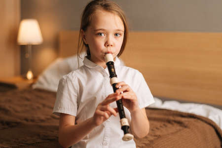 Close-up portrait of talented adorable little girl playing flute sitting on bed in bedroom, looking at camera. Child practices playing musical instrument at home.の写真素材