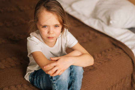 Top close-up view of distraught beautiful little girl with wet eyes from tears sits on bed in bedroom and looking at camera. Depressed child feeling pressure, thinking of kid problems.の写真素材
