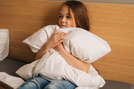 Happy adorable little girl sitting alone on bed hugging big pillow and looking away. Concept of leisure activity at home.の写真素材