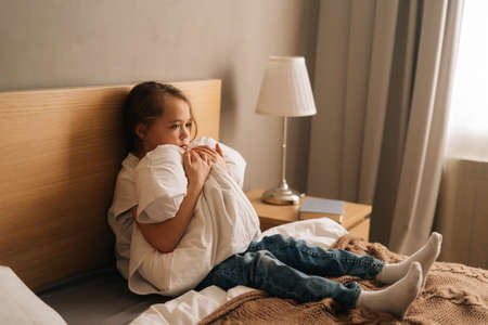 Upset little girl crying sitting alone in fetal position hiding face and rocking on bed hugging big pillow. Concept of child depression.の写真素材