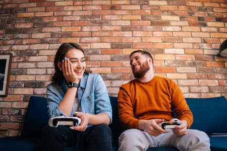 Front view of happy laughing young couple holding controllers and playing video games on console sitting together on couch at cozy living room. Concept of leisure activity of lovers at home.の写真素材