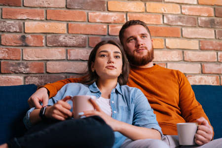 Portrait of young loving couple hugging, holding cups of warm drink and watching romantic movie on TV at evening together sitting on comfortable sofa in cozy living room. Concept of household life.の写真素材