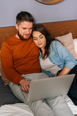 Cheerful Caucasian young couple watching video online on laptop lying in bed at night, cuddling and watching movie on computer. Concept of leisure activity of lovers at home.の写真素材