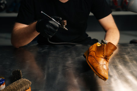 Close-up front view of unrecognizable shoemaker wearing black gloves spraying paint of light brown leather shoes. Concept of cobbler artisan repairing and restoration work in shoe repair shop.の写真素材