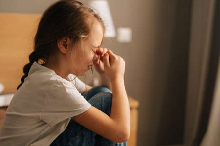 Close-up side view of upset little girl crying sitting alone on bed in bedroom. Unhappy child feeling pressure, thinking of kid problems. Concept of child depression.の写真素材
