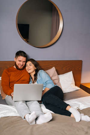 Cheerful Caucasian young couple watching video online on laptop lying in bed at night, cuddling and watching movie on computer. Concept of leisure activity of lovers at home.の写真素材