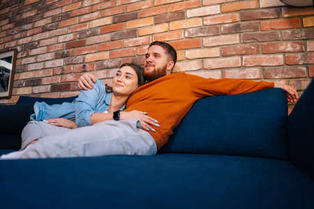Portrait of young hugging couple talking, holding cups of warm drink and watching romantic movie on TV together sitting on comfortable sofa at cozy living room. Concept of household life.の写真素材