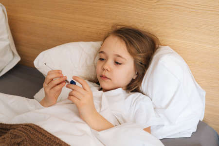 Close-up face of cute little sick girl with high fever lying under blanket and looking at...