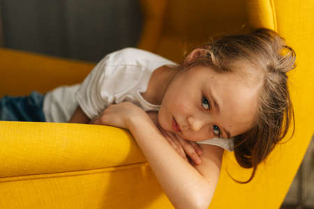 Close-up face of upset little girl with wet eyes from tears lying on yellow chair and looking at camera. Concept of child depression.の写真素材
