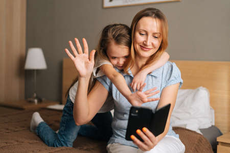 Happy young little girl embracing smiling mother greeting waving hands during online video call using mobile phone, looking on a display of smartphone. Concept of family leisure activity at home.の写真素材
