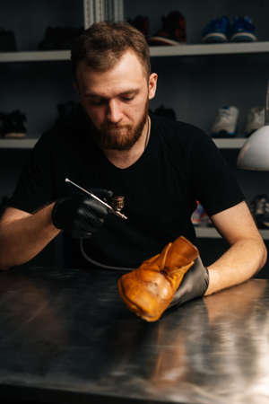 Front view of bearded shoemaker wearing black gloves spraying paint of light brown leather shoes, close-up. Concept of cobbler artisan repairing and restoration work in shoe repair shop.の写真素材