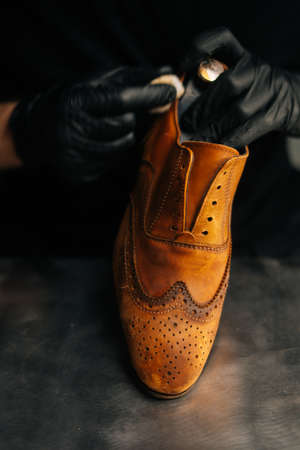 Close-up of shoemaker wearing black latex gloves cleaning old light brown leather shoes with rag for...