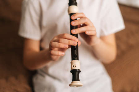 Close-up hands of talented adorable little girl playing flute sitting on bed in bedroom. Child practices playing musical instrument at home. Concept of leisure activity at home.の写真素材