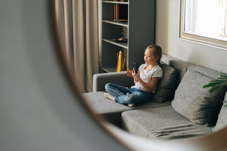 Mirror reflection of little girl talking on video call by mobile phone in living room sitting on cozy sofa. Adorable child looking fun video on cellphone sitting on couch.の写真素材