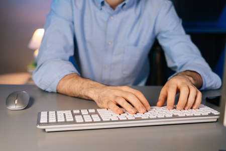 Close-up view of hands of unrecognizable man using wireless keyboard and computer mouse at dark room...