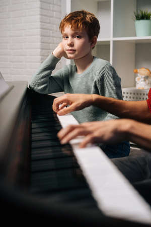 Focused redhead child student boy listening his teacher play the piano during lesson and looking at camera. Father teaching son to play musical instrument in living room. Concept of music education.の写真素材