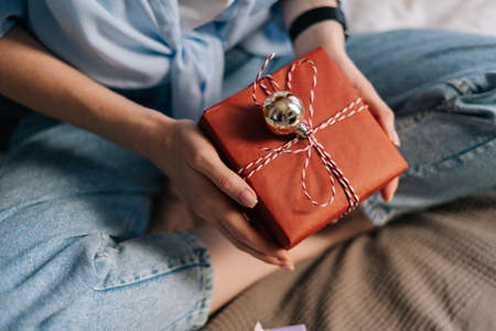 Close-up view of hands of unrecognizable woman holding gift box with holiday present sitting on lotus position at bed in light bedroom.の写真素材