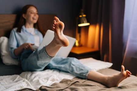 Cheerful redhead young woman chatting online via video call on laptop in bedroom lying at bed. Concept of leisure activity red-haired female at home during self-isolation.の写真素材