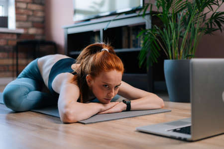 Sporty redhead young woman lying on floor and using laptop computer at apartment. Sporty smiling female having online chatting via notebook while resting after sport training.の写真素材