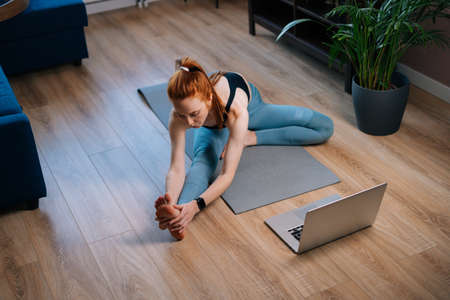 Top view of sporty redhead young woman working out, doing stretching exercise on yoga mat while watching fitness video online on laptop. Concept of sports training red-haired lady during quarantine.の写真素材