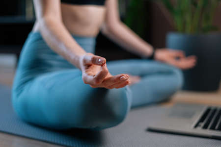 Close-up of unrecognizable young woman meditating at home sitting in lotus position on yoga mat near...