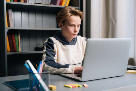 Close-up of serious pupil boy using typing laptop computer doing online lesson via Internet at desk, selective focus. Child schoolboy doing homework at home. Concept of remote distance education.の写真素材
