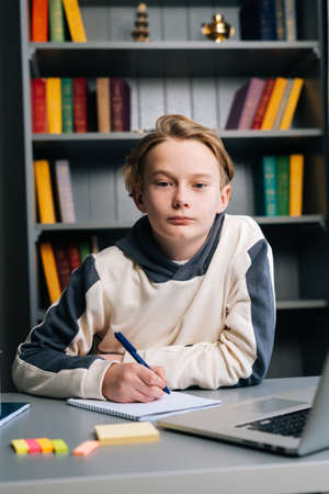 Close-up portrait of pupil boy making notes in notebook with pen sitting at desk with laptop computer, looking at camera. Child schoolboy doing homework at home. Concept of remote online education.の写真素材