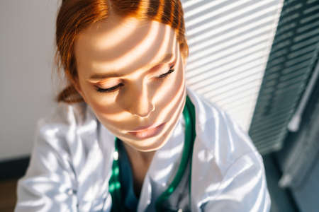 Close-up face of desperated sad young female doctor in white coat sitting on floor hugging legs with hands near window. Unhappy woman physician depression feeling worried about malpractice.の写真素材