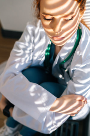 Close-up face of desperated sad young female doctor in white coat sitting on floor hugging legs near window. Unhappy woman physician depression feeling worried about professional malpractice.の写真素材