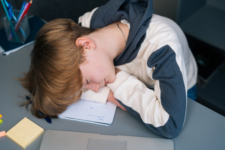 Close-up top view of tired pupil boy sleeping on copybook in front of laptop after online lesson via...