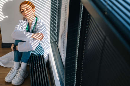 Close-up of frustrated sad young female doctor in white coat sitting on floor hugging legs with hands near window. Unhappy woman physician depression feeling worried about professional malpractice.の写真素材