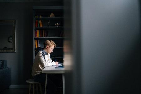 Side view of focused pupil boy making notes in copybook and using laptop computer sitting at desk near window. Child schoolboy doing homework at home. Concept of remote online distance education.の写真素材