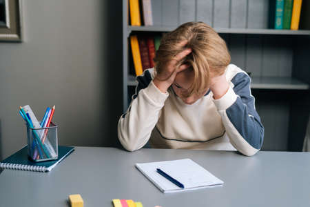 Portrait of exhausted pupil boy tired from studying holding head head with hands while sitting at desk with workbook. Child schoolboy doing homework at home.の写真素材