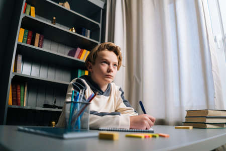 Low-angle view of thinking pupil boy thoughtful writing in notebook with pen sitting at desk near window. Close-up of child schoolboy doing homework at home.の写真素材