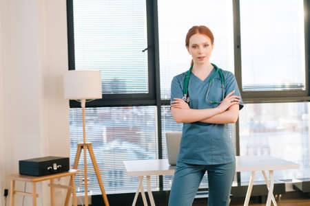 Portrait of serious female doctor in blue green uniform standing arms crossed near window in sunny day in medical clinic office. Redhead woman surgeon posing with stethoscope looking at camera.の写真素材