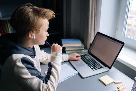 Top back view of pupil boy wearing wireless earphone learning online using laptop making notes in copybook sitting at desk at home near window. Concept of distance education.の写真素材