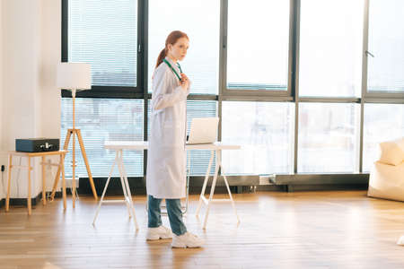 Portrait of serious female doctor in white coat standing on background of window in sunny day in light medical clinic office. Young redhead woman physician posing with stethoscope looking away.の写真素材